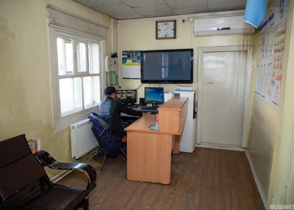 Ширээн дээр суугаад жижиг оффист компьютер дээр бичиж байгаа хүн. Өрөө нь цайвар шар ханатай, ханын цаг, ширээний дээгүүр зурагт дэлгэцтэй, хөшигтэй цонхтой. Өрөөний эргэн тойронд зурагт хуудас, оффисын тоног төхөөрөмж харагдаж байна. - Business.MN