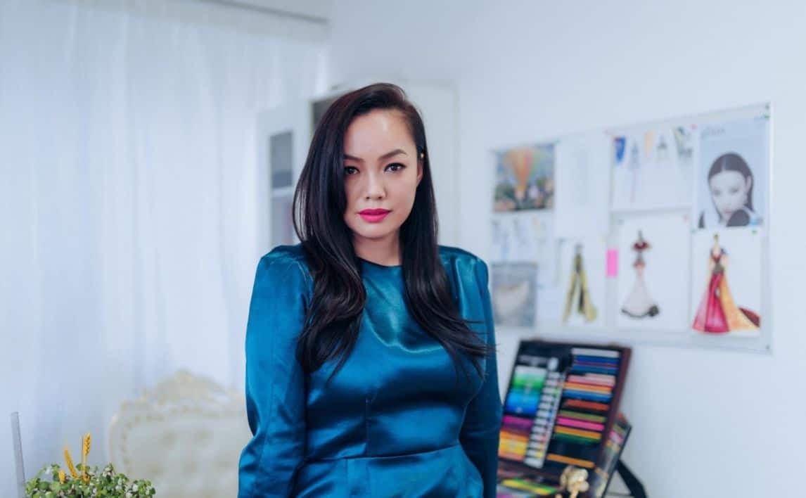 Гялалзсан цайвар өнгийн даашинз өмссөн урт хар үстэй эмэгтэй цагаан хөшигтэй өрөөнд зогсож байна. Түүний ард өнгөт харандаа бүхий тавиур, янз бүрийн зураг, хийц бүхий хана байгаа нь ажил хэрэгч, бүтээлч ажлын байрны уур амьсгалыг илэрхийлдэг. - Business.MN