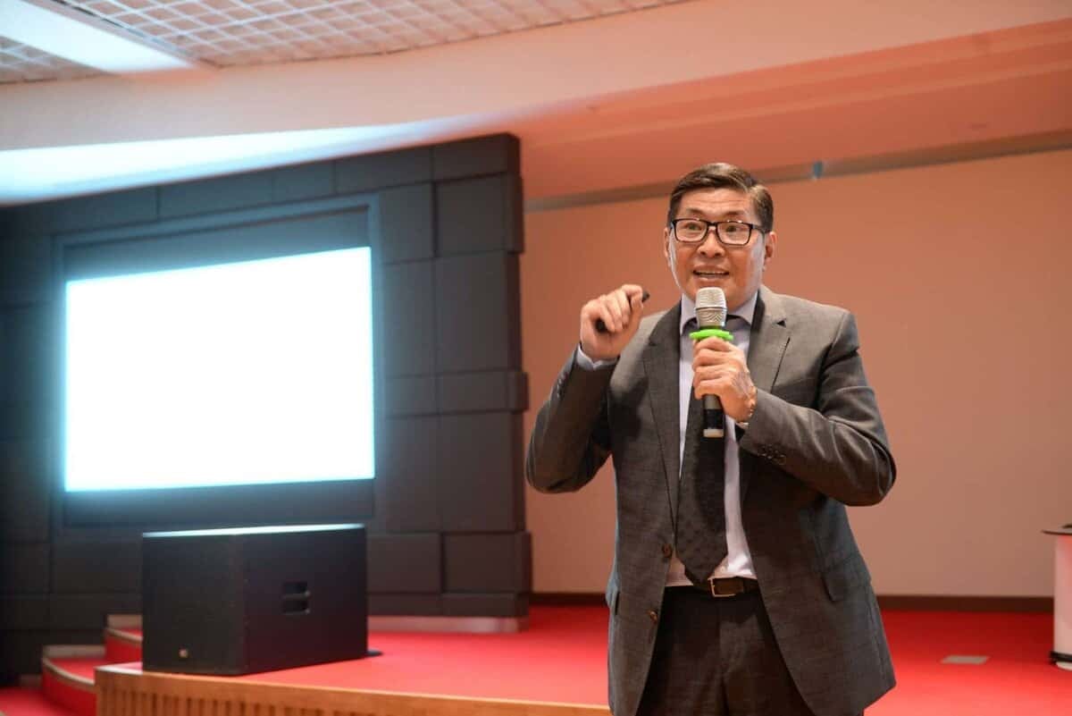 Харанхуй костюм, зангиа зүүсэн эр улаан хивсэнцэртэй тайзан дээр зогсож байхдаа микрофонд ярьж байна. Тэр нөгөө гараараа дохиж байна. Цаана нь цагаан гэрэлтэй том дэлгэц байгаа нь бизнесийн арга хэмжээ зохион байгуулах боломжтой танхим эсвэл хурлын танхим болохыг харуулж байна. - Business.MN