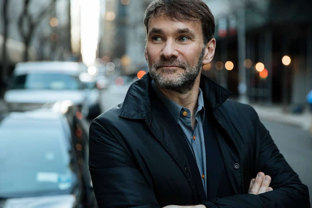 A man with a beard and short hair stands on a city street with his arms crossed. He is wearing a dark jacket and gazing off to the side. The background is slightly blurred, showing cars and buildings.