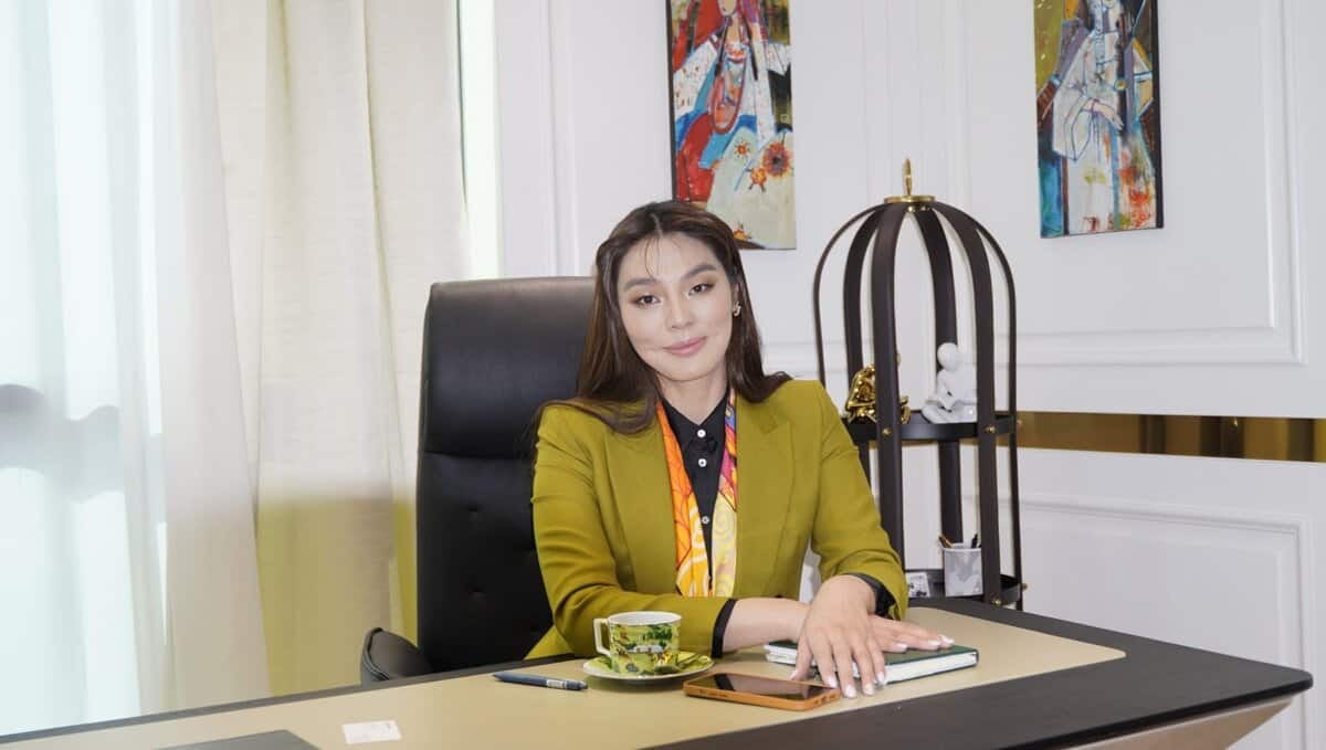 Ногоон пиджак өмссөн бизнесмэн эмэгтэй аяга цай, ухаалаг утас, номтой ширээний ард сууж байна. Тэрээр ханан дээрх өнгө өнгийн зураг, гоёл чимэглэлийн шувууны тор зэргийг багтаасан орчин үеийн чимэглэлтэй оффист. Энтрепренер камерын өмнө үл ялиг инээмсэглэнэ. - Business.MN