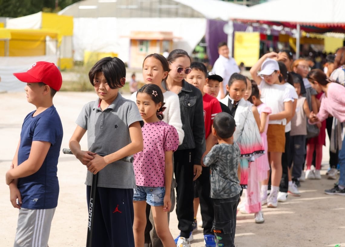 A group of children stand in line outdoors, waiting for an activity. Some wear hats and sunglasses. Tents and colorful booths are visible in the background on a sunny day.