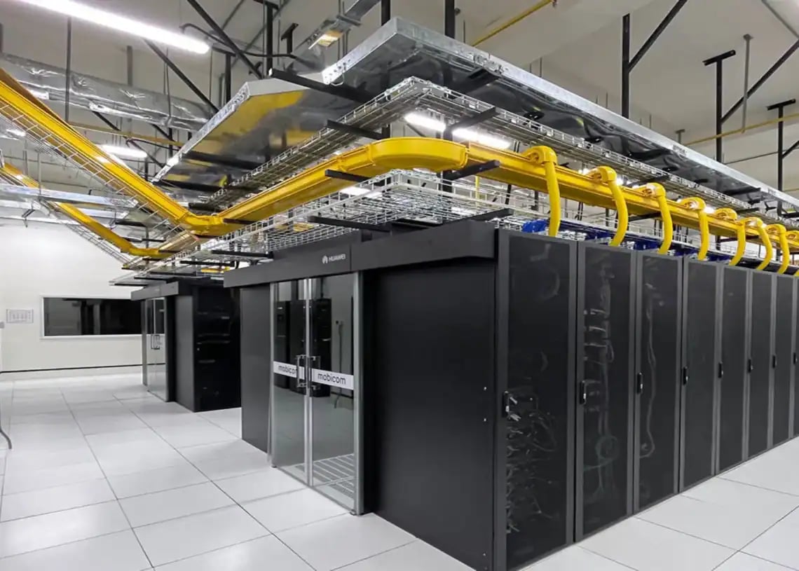 A modern data center with rows of black server racks. Yellow cable trays run across the ceiling. The room has a white tiled floor and exposed structural elements on the ceiling.