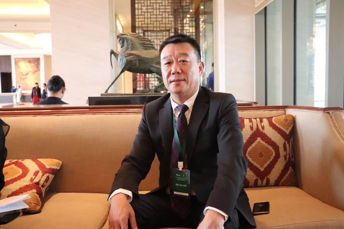 Орчин үеийн лобби дахь шаргал буйдан дээр костюм өмссөн хүн сууж байна. Тэрээр хүзүүндээ хурлын тэмдэг зүүж, нэг гараа буйдан дээр тавьдаг. Ар талд нь гоёл чимэглэлийн баримал, хийсвэр урлагийн бүтээлүүд харагдаж байна. - Business.MN