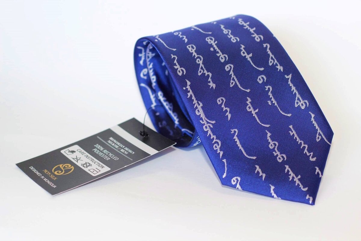 A blue necktie with white script in a foreign language, displayed against a white background. The tie has a black and white tag attached to it showing brand and care information.