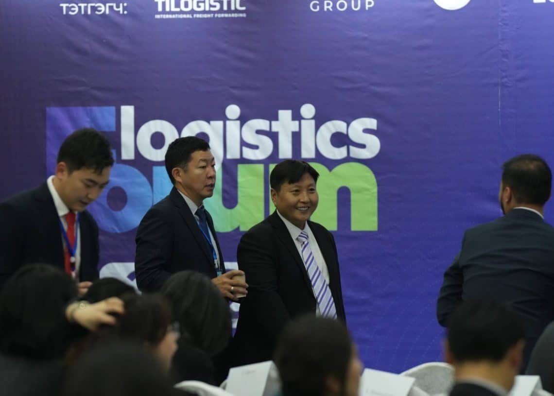 A group of people in formal attire at the Монголын Тээвэр Зуучлагчдын Нэгдсэн Холбоо’s "Ложистикс Форум 2024" event. The backdrop displays "logistics forum." Some attendees are seated, while others stand and engage in conversation.