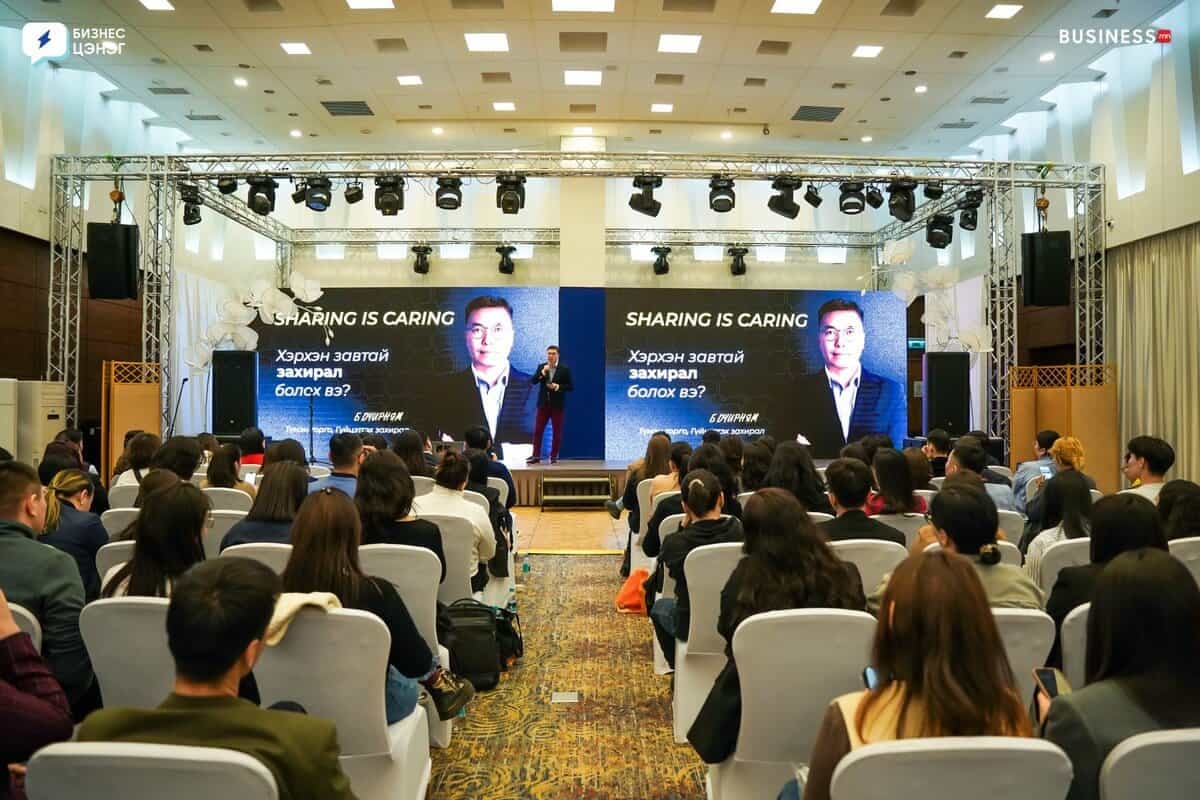 A person speaks on stage in a conference room with a large audience seated. The screen behind displays "SHARING IS CARING" with an image of another person. Attendees face forward, listening intently. The room is well-lit with a patterned carpet.