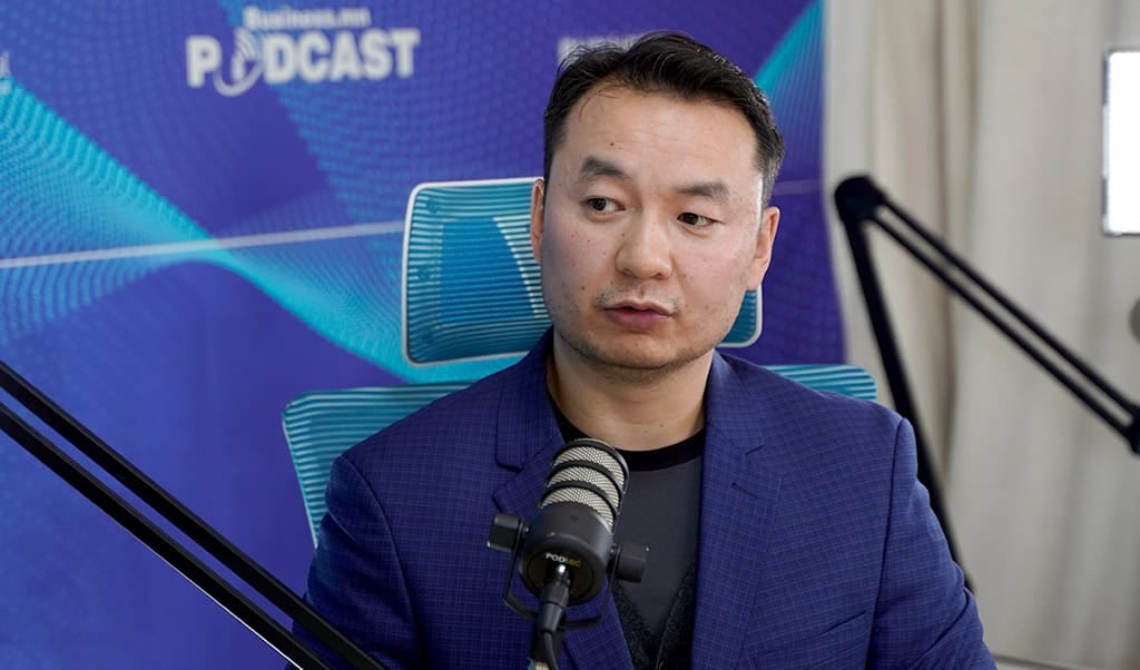 Цэнхэр пиджак, хар цамц өмссөн хүн түүний өмнө микрофоноор ярьж байна. Тэрбээр цэнхэр, цагаан долгионы хээтэй "PODCAST" гэсэн дэвсгэртэй цэнхэр сандал дээр сууж байна. Хоёр микрофоны тавцан харагдаж байгаа бөгөөд хэлэлцүүлэг нь компанийг удирдаж буй энтрепренерийн бизнесийн стратегийн талаар эргэлддэг. - Business.MN