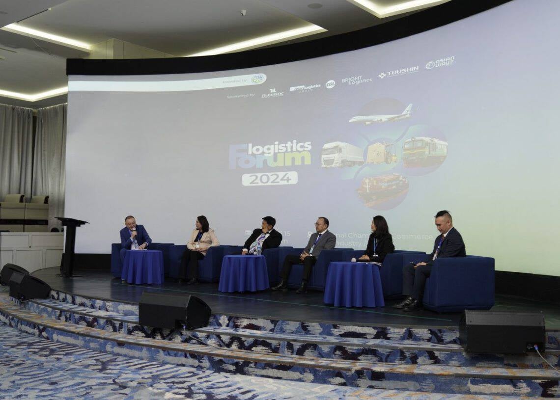 Six panelists sit on stage in front of a large screen displaying "Монголын Тээвэр Зуучлагчдын Нэгдсэн Холбоо 'Ложистикс Форум 2024’." Seated in blue chairs, they engage deeply in discussion within a room boasting a modern design and light-colored carpet.