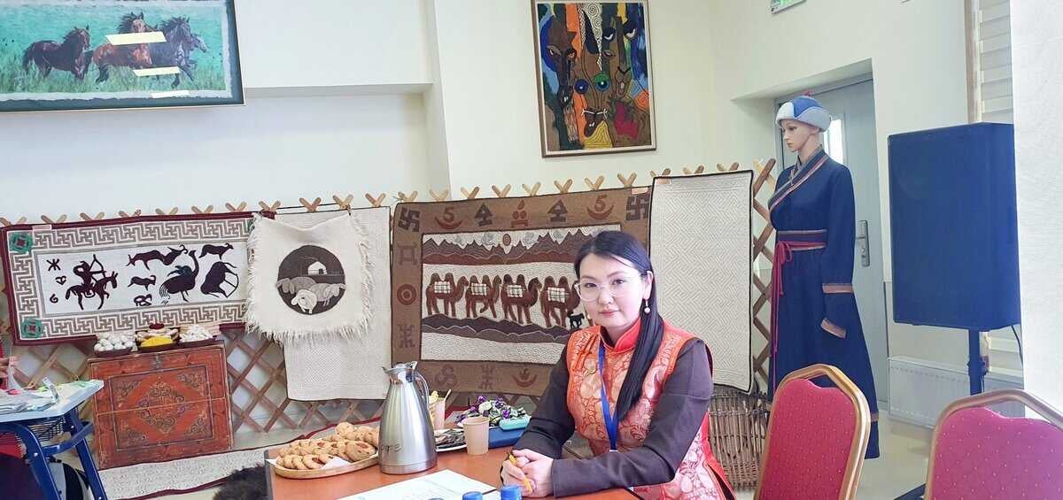 A woman in traditional attire, embodying Үе дамжсан уран шаглаа, sits at a table with a thermos and snacks. Behind her, tapestries depicting horses and other designs fill the space. A mannequin dressed in similar traditional clothing adds to the scene's timeless ambiance.