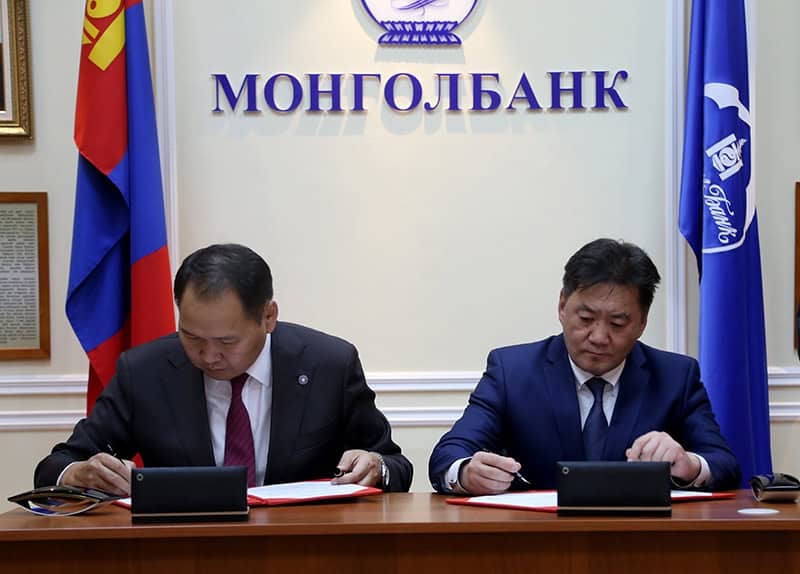 Костюм өмссөн хоёр эрэгтэй ширээний ард суугаад бичиг баримтад гарын үсэг зурж байна. Тэдний ард Монголбанкны (“Монголбанк”) бэлгэ тэмдэг, нэрийг цэнхэр үсгээр бичсэн хана бий. Сүлд тэмдгийн хоёр талд тугнууд байрлуулсан нь ирээдүйн эдийн засгийн хүчин чармайлтыг тодорхойлох бизнесийн чухал түншлэлийн бэлгэдэл юм. - Business.MN