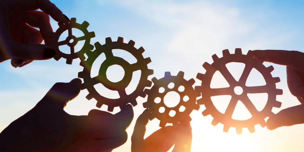Four hands hold interlocking gears against a bright sky, symbolizing teamwork, collaboration, and connection as sunlight shines through the gear spokes.