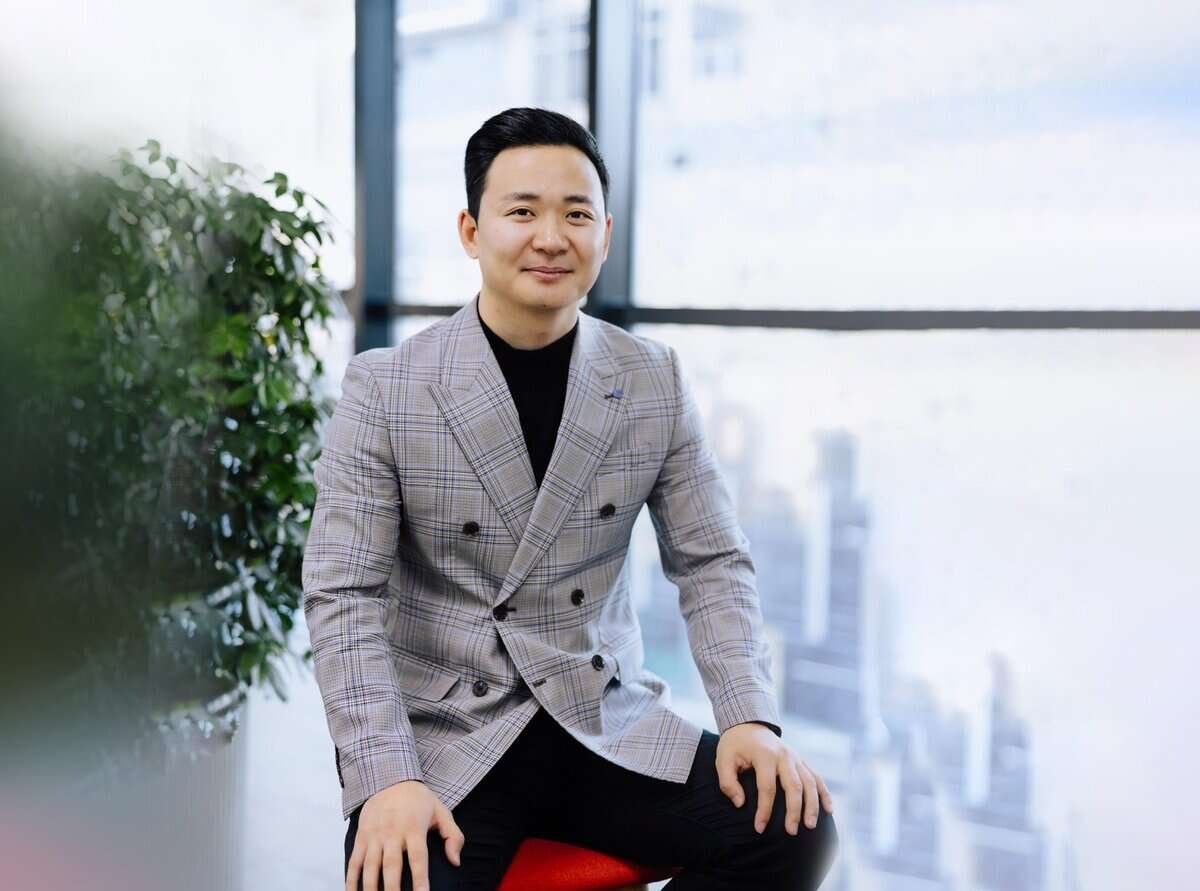 Саарал алаг давхар дээлтэй пиджак, хар өмд өмссөн эрэгтэй сууж, инээмсэглэж байна. Дотор, том цонхтой, арын хэсэг ногоон байгууламжтай энтрепренер өөртөө итгэлтэй байж, дараагийн бизнесийнхээ талаар бодож байгаа байх. - Business.MN