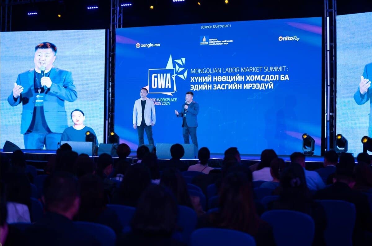 Хоёр илтгэгч тайзан дээр зогсож, 2024 оны Монголын хөдөлмөрийн зах зээлийн дээд хэмжээний уулзалтад илтгэл тавьж байна. Арын дэвсгэр дээр GWA лого бүхий үйл явдлын брэндийг харуулдаг. Үзэгчид суудалдаа сууж, хүний нөөцийн менежментийн шинэчлэлийг хэлэлцэж байгааг анхааралтай ажиглаж байна. - Business.MN