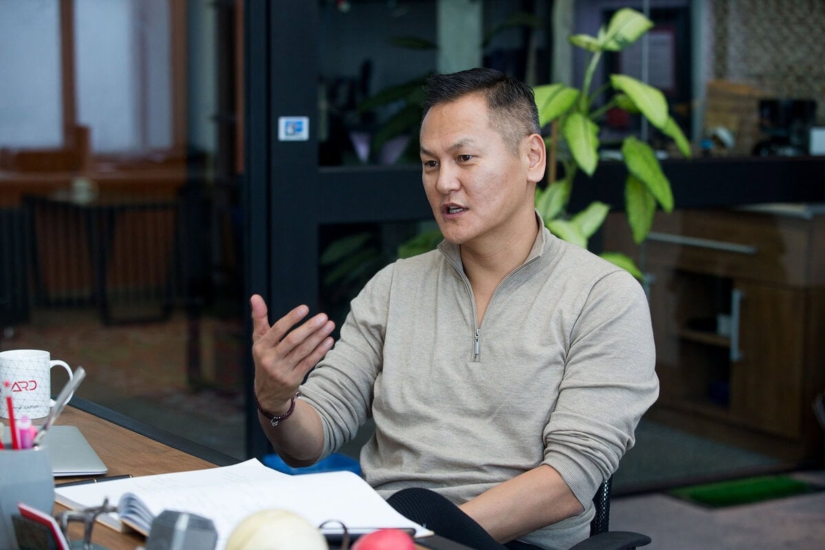 Энгийн хувцас өмссөн хүн ширээний ард сууж, ярьж байхдаа дохиж, ширээн дээр нээлттэй ном, оффисын эд зүйлс тавьсан нь бизнесийн идэвхтэй хэлэлцүүлэгт оролцож буй энтрепренерийн сүнсийг илэрхийлдэг. - Business.MN