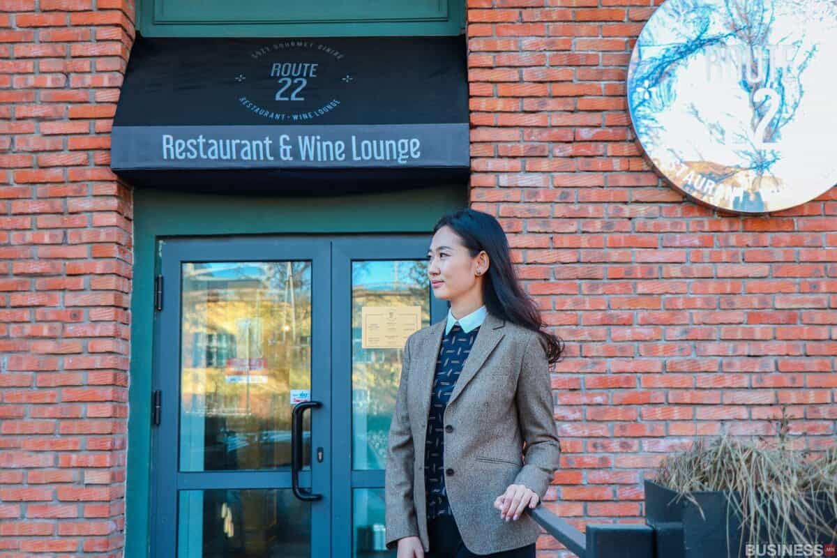 Хүрэн пиджак өмссөн эмэгтэй "Route 22 Restaurant & Wine Lounge" гэсэн бичигтэй тоосгон барилгын гадаа зогсож байгаа нь бизнесээ өргөжүүлэхэд бэлэн бизнес эрхлэгчийн өөртөө итгэлтэй байдлыг илтгэнэ. - Business.MN