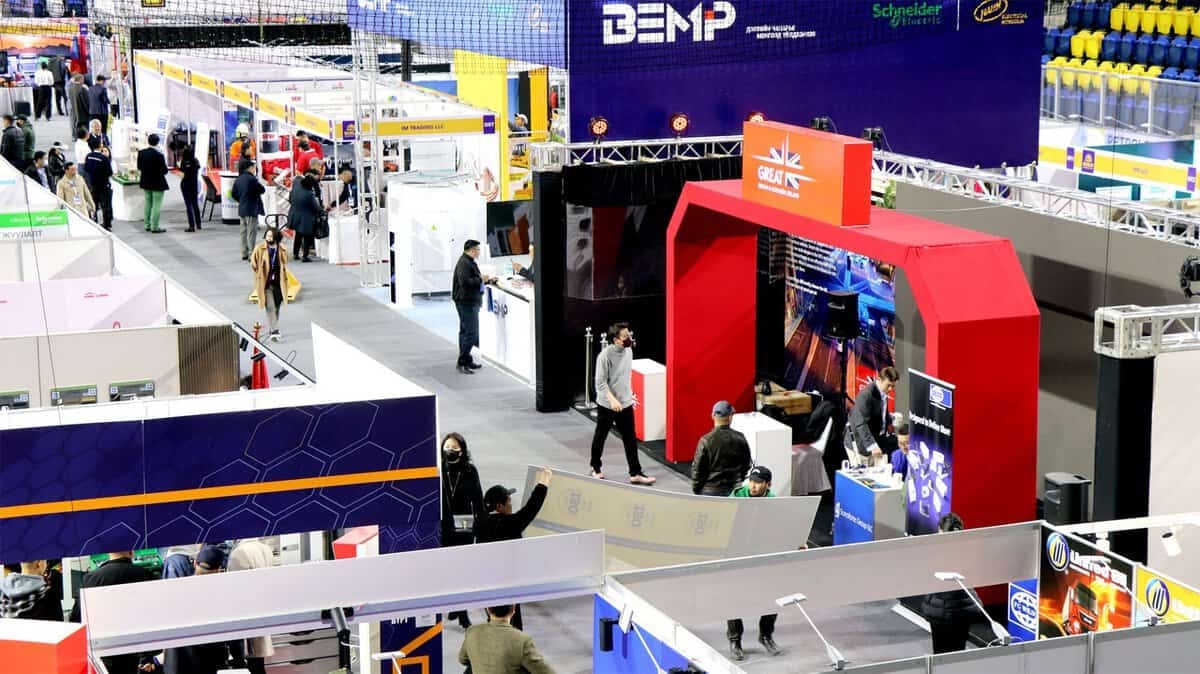 Олон тооны өнгөлөг лангуу, үзмэр бүхий хөл хөдөлгөөн ихтэй үзэсгэлэнгийн танхим. Оролцогчид, тэдний ихэнх нь энтрепренерүүд, эргэн тойрон алхаж, янз бүрийн үзэсгэлэнд оролцогчидтой уулзаж байна. Янз бүрийн компани, бүтээгдэхүүнийг онцлон харуулсан баннер, тэмдэгтүүд харагдаж байна. Уур амьсгал нь эрч хүчтэй, эрч хүчтэй байдаг. - Business.MN