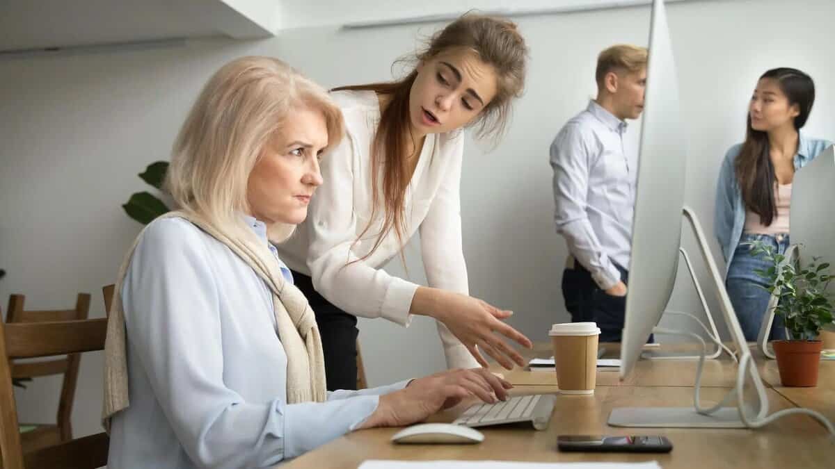 Нэгэн залуу эмэгтэй ширээний ард компьютерийн ард сууж буй хөгшин эмэгтэйтэй ярилцахаар бөхийж байна. Хөгшин эмэгтэй, магадгүй энтрепренер, төвлөрсөн илэрхийлэлтэй байдаг. Цаана нь эрэгтэй, эмэгтэй хоёр зогсож, ярилцаж байна. Ширээн дээр ургамал, кофены аяга байна. - Business.MN