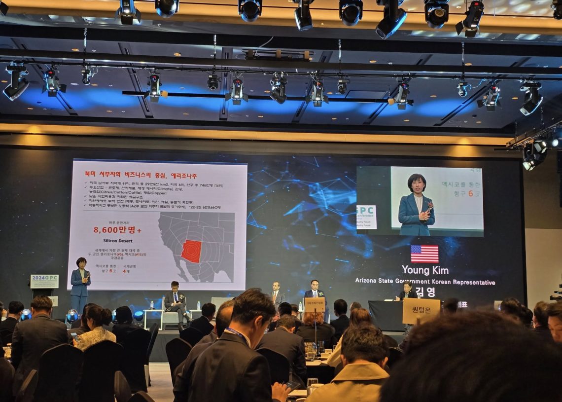 A conference room filled with attendees. A large screen displays a presentation slide with a map and text. Four people are on stage, and a speaker is presenting. The room is lit with ceiling lights.