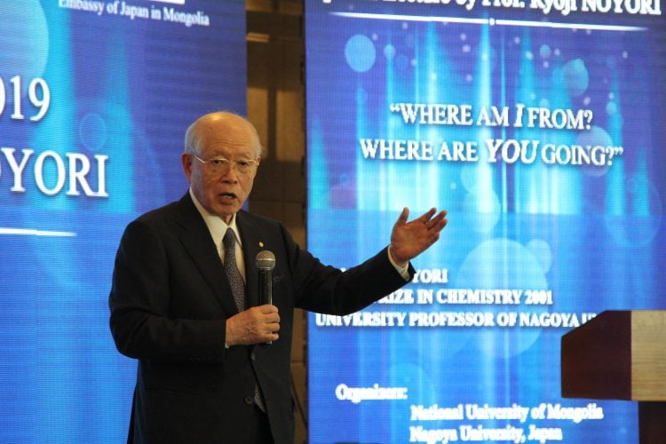 Костюм өмссөн өндөр настан тайзан дээр зогсож, микрофоноор ярьж байна. Түүний ард байгаа проекц дээр "БИ ХААНА БАЙНА? ЧИ ХААЧИН ЯВЖ БАЙГАА ЮМ?" болон үйл явдлын дэлгэрэнгүй мэдээлэл. - Business.MN