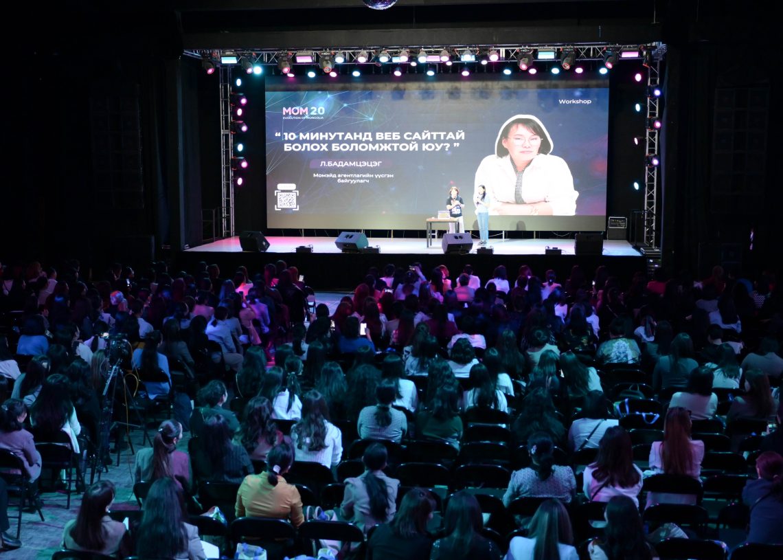 A large audience sits in a dimly lit auditorium facing a stage. A speaker, seated, presents a slideshow with text and an image on a screen. The event features stage lighting and a large black backdrop.