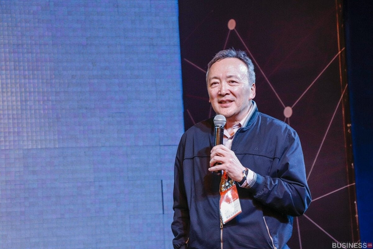 Хар хүрэмтэй эр тайзан дээр ярьж байхдаа микрофон барьж байна. Арын дэвсгэр нь том дэлгэц, геометрийн хэв маягтай. Иргэний үнэмлэхтэй оосор зүүдэг. - Business.MN