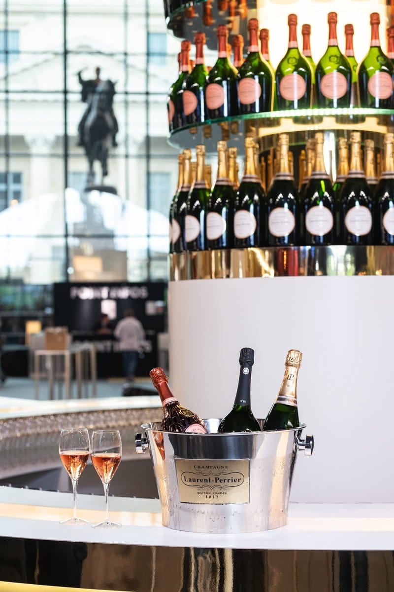 Ice bucket with three bottles of champagne and two filled flutes on a table. Behind them, shelves display multiple bottles of champagne. A silhouette of a horse statue is visible through large windows in the background.