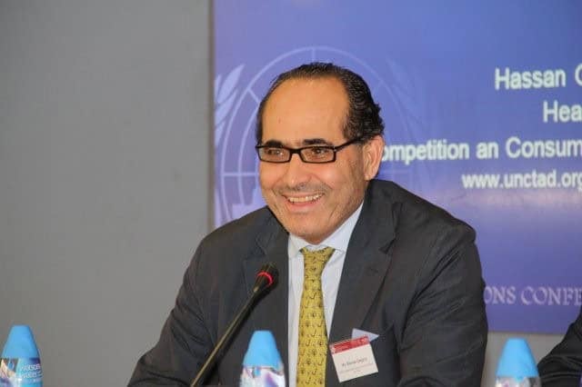 Костюм, зангиа зүүсэн эр микрофонтой ширээний ард инээмсэглэн сууж байна. Түүний ард цэнхэр дэвсгэр дээр текст болон вэбсайтын URL-г харуулдаг. Ширээн дээр хоёр савтай усны сав байна. - Business.MN