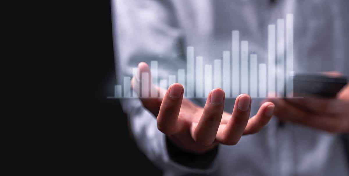 Бүдгэрсэн саарал цамцтай хүн гараа сунгаж, дээр нь голограмм 3D зураасан график байрлуулна. Арын дэвсгэр нь бараан өнгөтэй, нөгөө гар нь ухаалаг утас барьдаг. - Business.MN