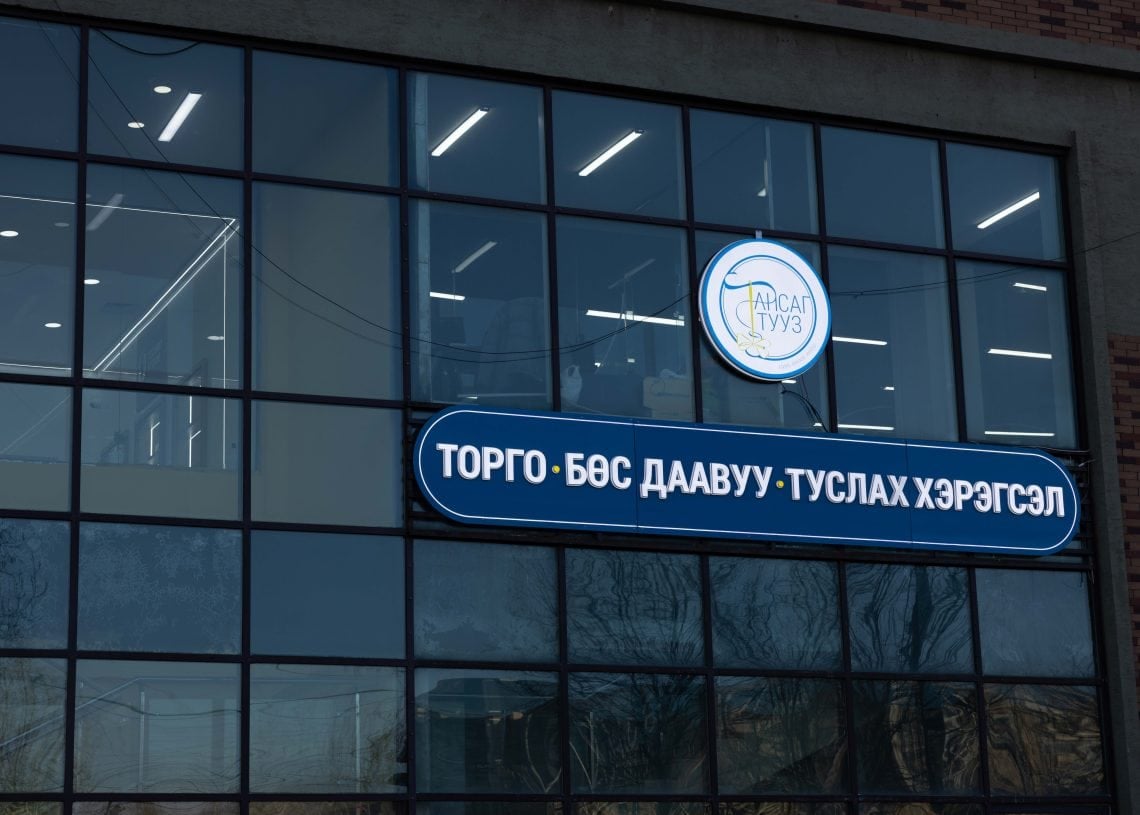 The image shows a building facade with large glass windows. Two illuminated signs are displayed, featuring text in a non-Latin script. Reflections of trees and the sky can be seen in the windows.