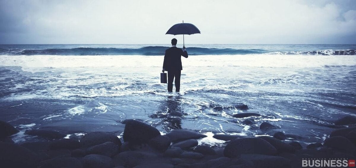 Костюм өмссөн хүн чулуурхаг эрэг дээр, далай руу харсан, шүхэр, цүнх барьсан зогсож байна. Тэнгэр бүрхэг, давалгаа эрэг рүү ойртож, гайхалтай, эргэцүүлэн бодох дүр зургийг бий болгодог. - Business.MN