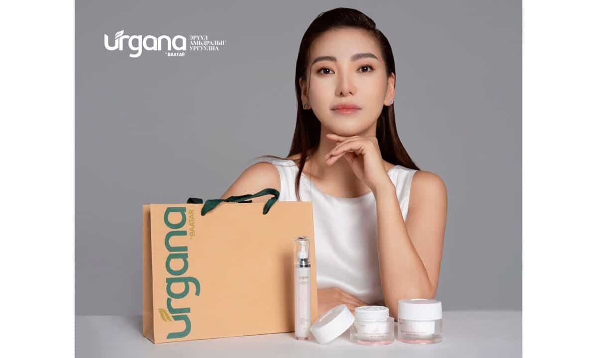 Урт хар үстэй, ханцуйгүй цагаан цамц өмссөн эмэгтэй ширээний ард суугаад гараа эрүүнийхээ доор тавив. Түүний өмнө "Ургана" гэсэн хэвлэсэн шаргал цаасан уут, арьс арчилгааны хэд хэдэн бүтээгдэхүүн байна. Арын дэвсгэр нь саарал өнгөтэй бөгөөд гоо сайхны салбарын энтрепренерт тохирсон дэгжин байдлыг илтгэнэ. - Business.MN
