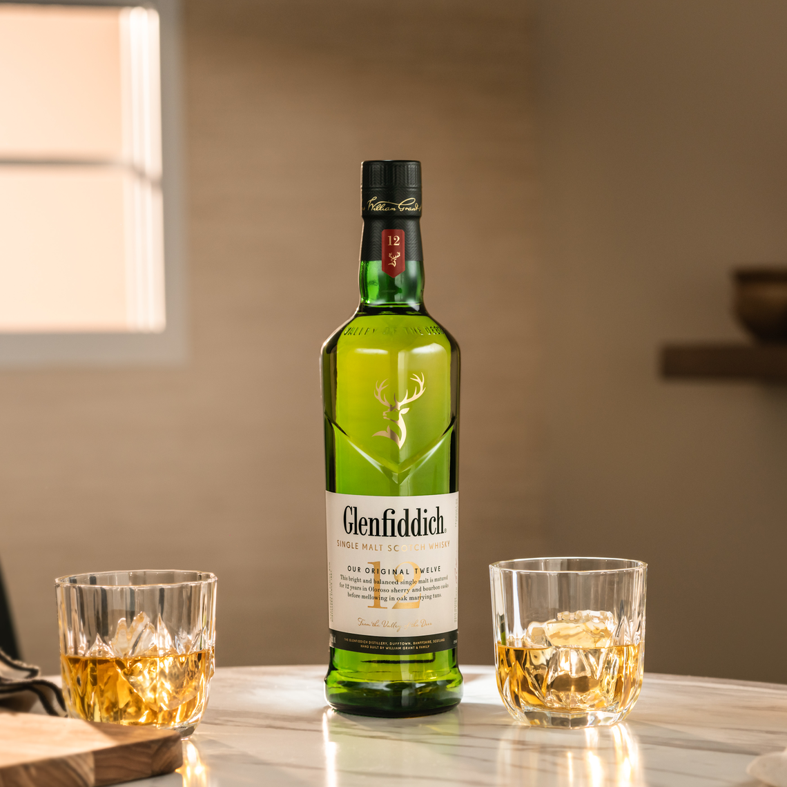 A bottle of Glenfiddich single malt Scotch whisky is on a table with two glasses filled with whisky and ice cubes. The background is softly lit, with a window and a blurred wall.