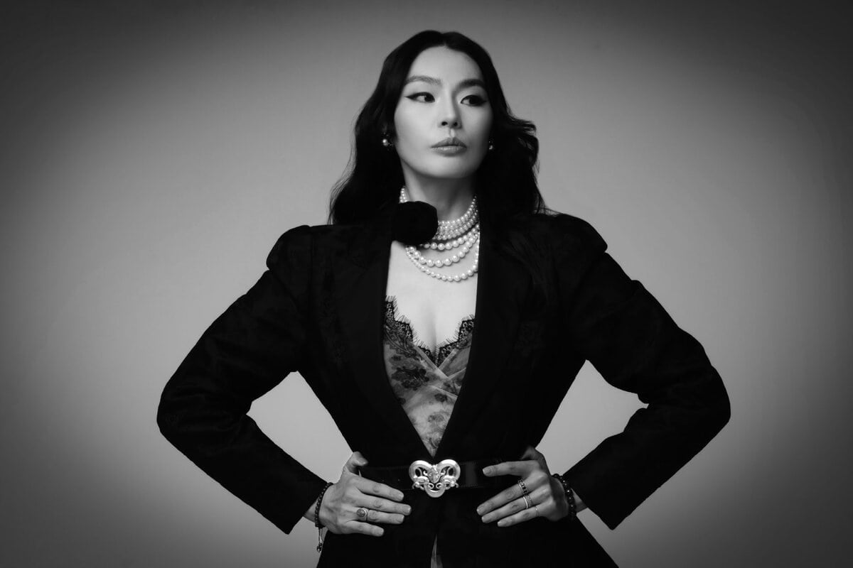 Хар нэхсэн тортой хувцас, сувдан хүзүүний зүүлттэй өөртөө итгэлтэй эмэгтэй гараа ташаандаа бариад, бүстэй хүрэм өмсөж, хар цагаан гэрэл зургийн энгийн дэвсгэр дээр хажуу тийшээ харж байна. - Business.MN