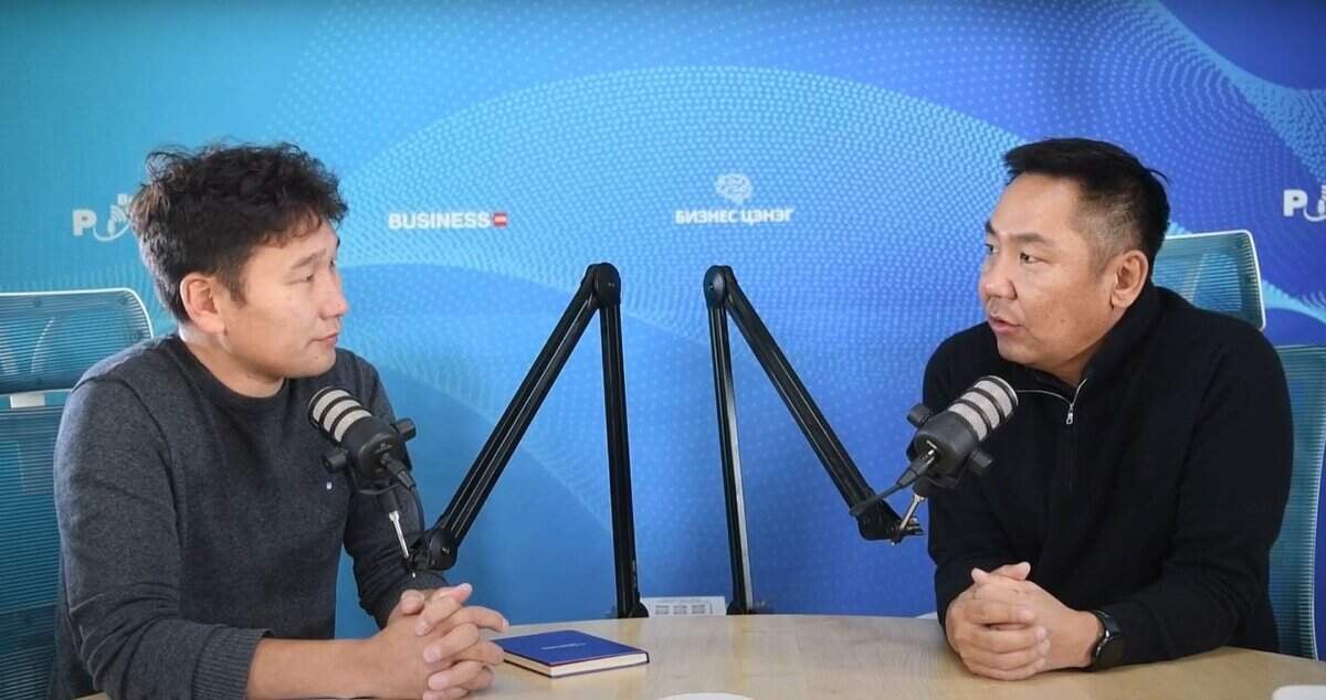 Хоёр эрэгтэй урд нь микрофонтой ширээний ард суугаад харилцан яриа өрнүүлж байна. Хоёулаа бараан өнгийн ноосон цамц өмссөн ба арын дэвсгэр дээр "BUSINESS CONUNDRY" болон "PURU" лого бүхий технологийн сэдэвтэй цэнхэр загвартай. Тэд ямар ч энтрепренертэй холбоотой бизнесийн стратегийн талаар ярилцаж байх үед дэвтэр ширээн дээр байрладаг. - Business.MN