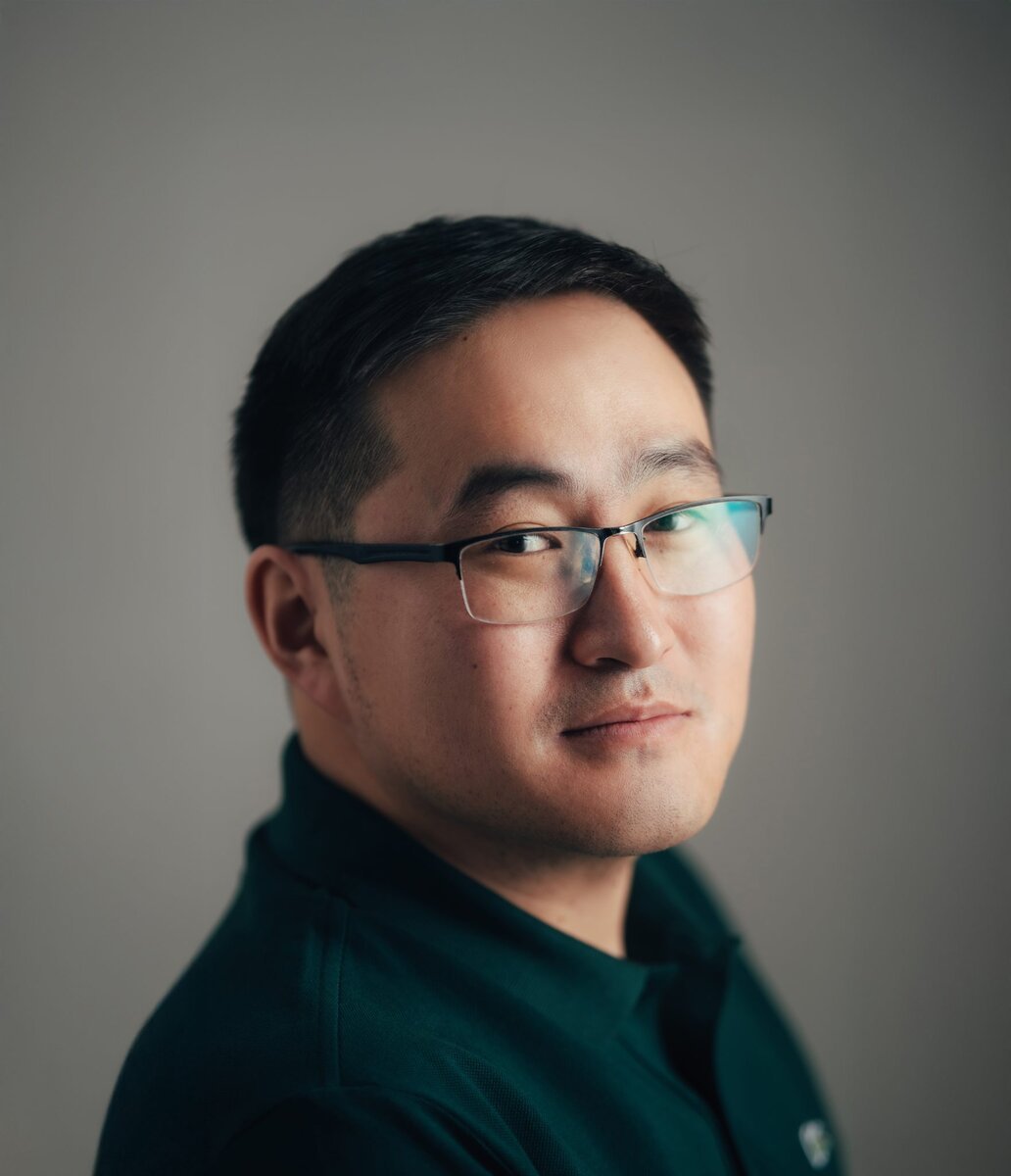 Man wearing glasses and a dark green shirt, looking at the camera with a neutral expression against a plain gray background.