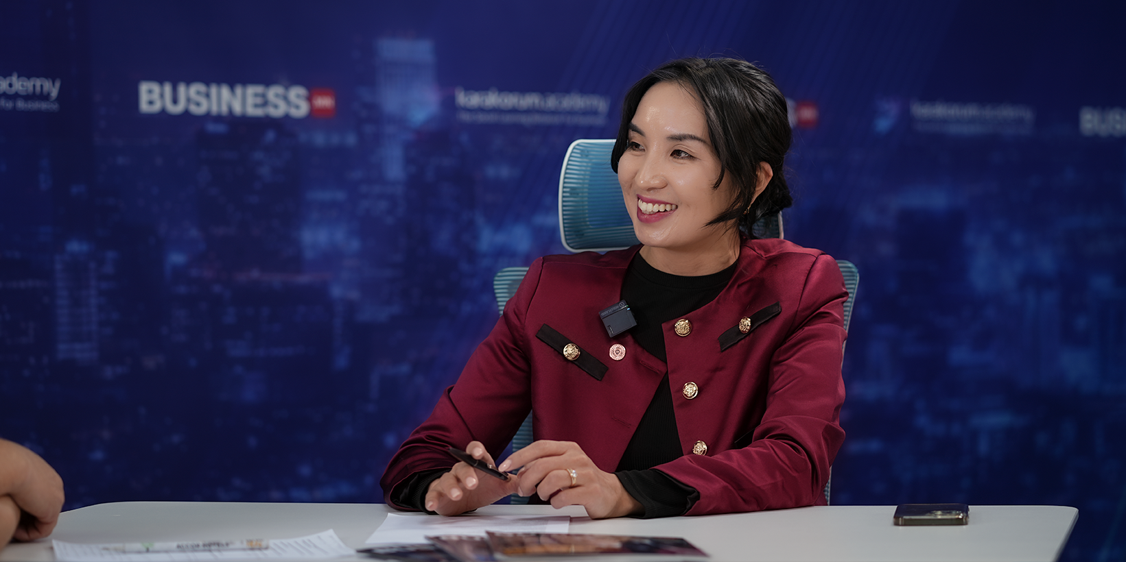 Алтан товчтой улаан пиджак өмссөн эмэгтэй ширээний ард суугаад инээмсэглэн үзэг барьжээ. Түүний урд талын ширээн дээр цааснууд байна. Тэр хувцсандаа микрофон хавчуулсан байна. Арын дэвсгэр дээр "БИЗНЕС" гэсэн үг харагдах бөгөөд түүний энтрепренерийн үүргийг онцлон харуулсан корпорацийн орчин мэт харагдана. - Business.MN