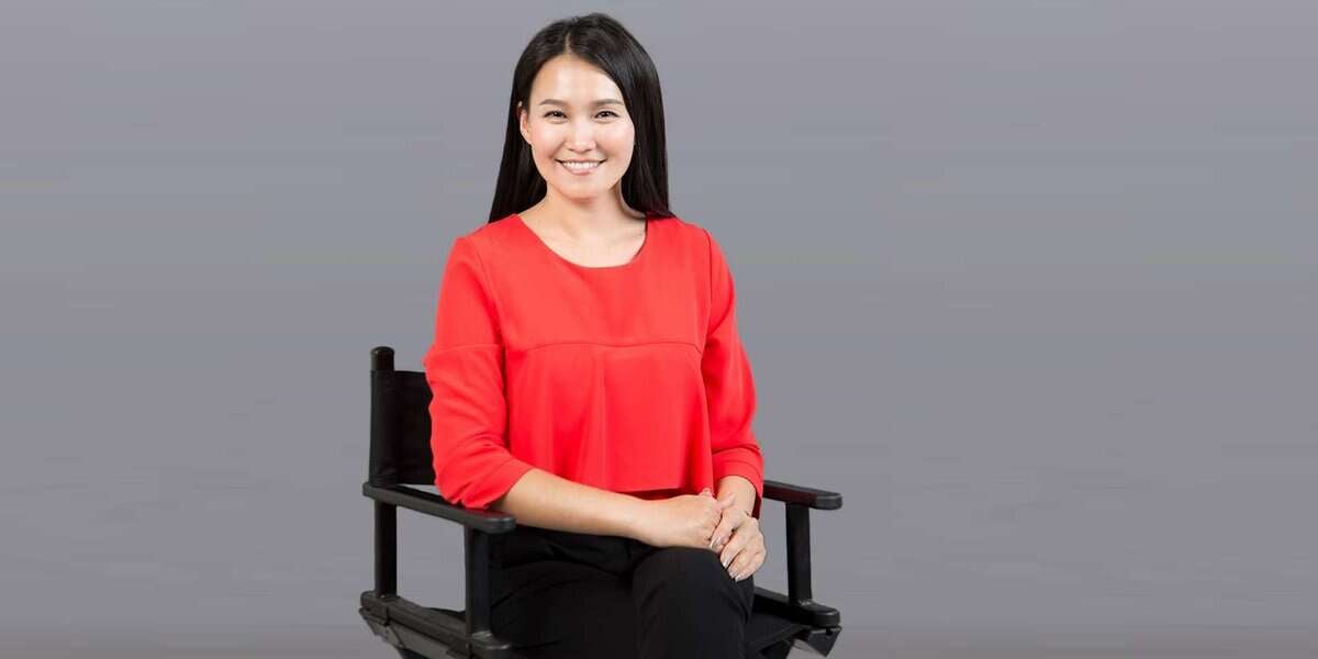 Улаан цамц, хар өмд өмссөн урт хар үстэй хүн хар сандал дээр инээмсэглэн сууж, камер руу харна. Арын дэвсгэр нь энгийн саарал өнгөтэй. - Business.MN