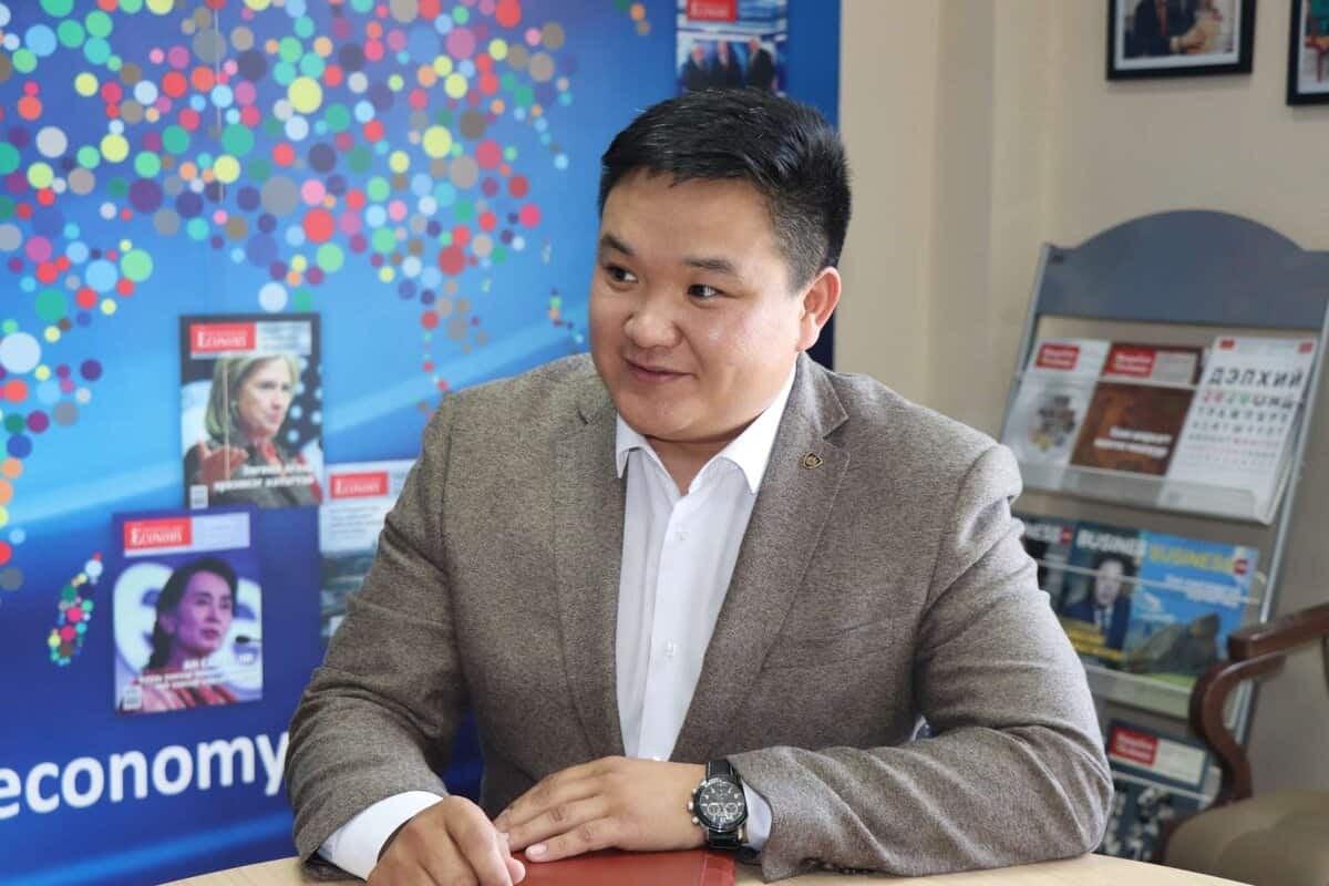 Бор пиджак өмссөн эрэгтэй, магадгүй энтрепренер, өнгө өнгийн сэтгүүл, цаана нь цэнхэр өнгийн зурагт хуудас бүхий ширээний ард сууж байна. - Business.MN