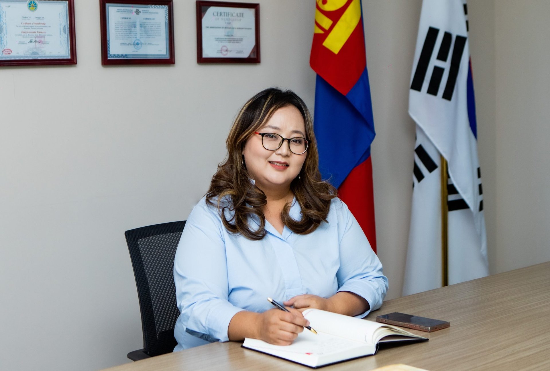 A woman in a light blue blouse is seated at a desk, smiling, with an open notebook and pen in hand. Behind her are framed certificates and flags.