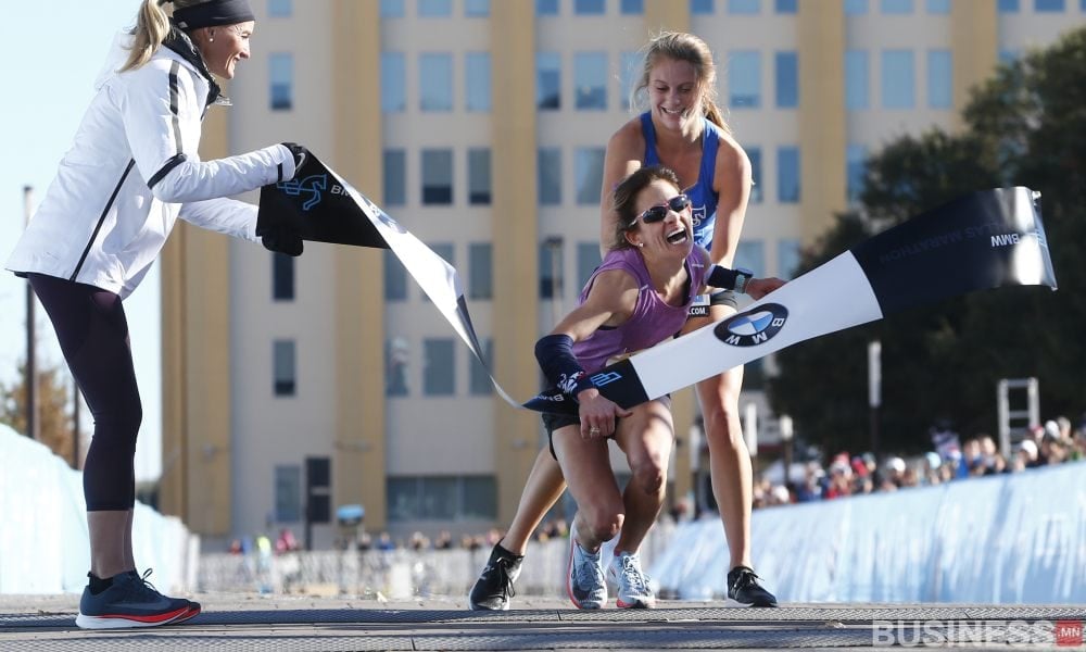 Нил ягаан өнгийн хувцастай гүйгч барианы туузыг том инээмсэглэлээр таслав, түүнд цэнхэр цамц өмссөн өөр хүн туслав. Цагаан хүрэмтэй гуравдагч хүн туузыг барьдаг бөгөөд цаана нь барилга харагдаж байна. - Business.MN