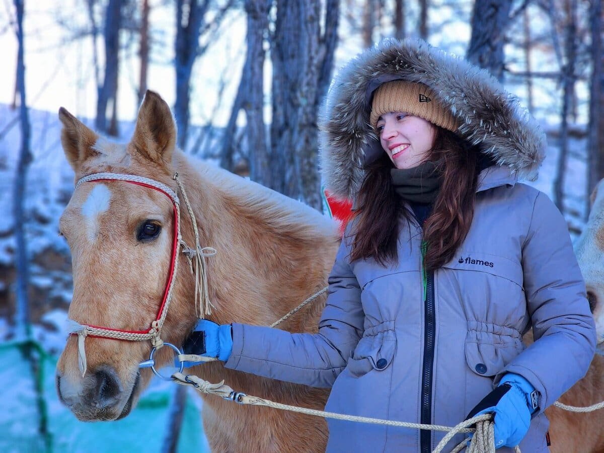 A woman dressed in a brown fur-lined hooded jacket, beanie, and blue gloves stands outdoors in snowy woods, smiling while holding the reins of a light brown horse beside her.