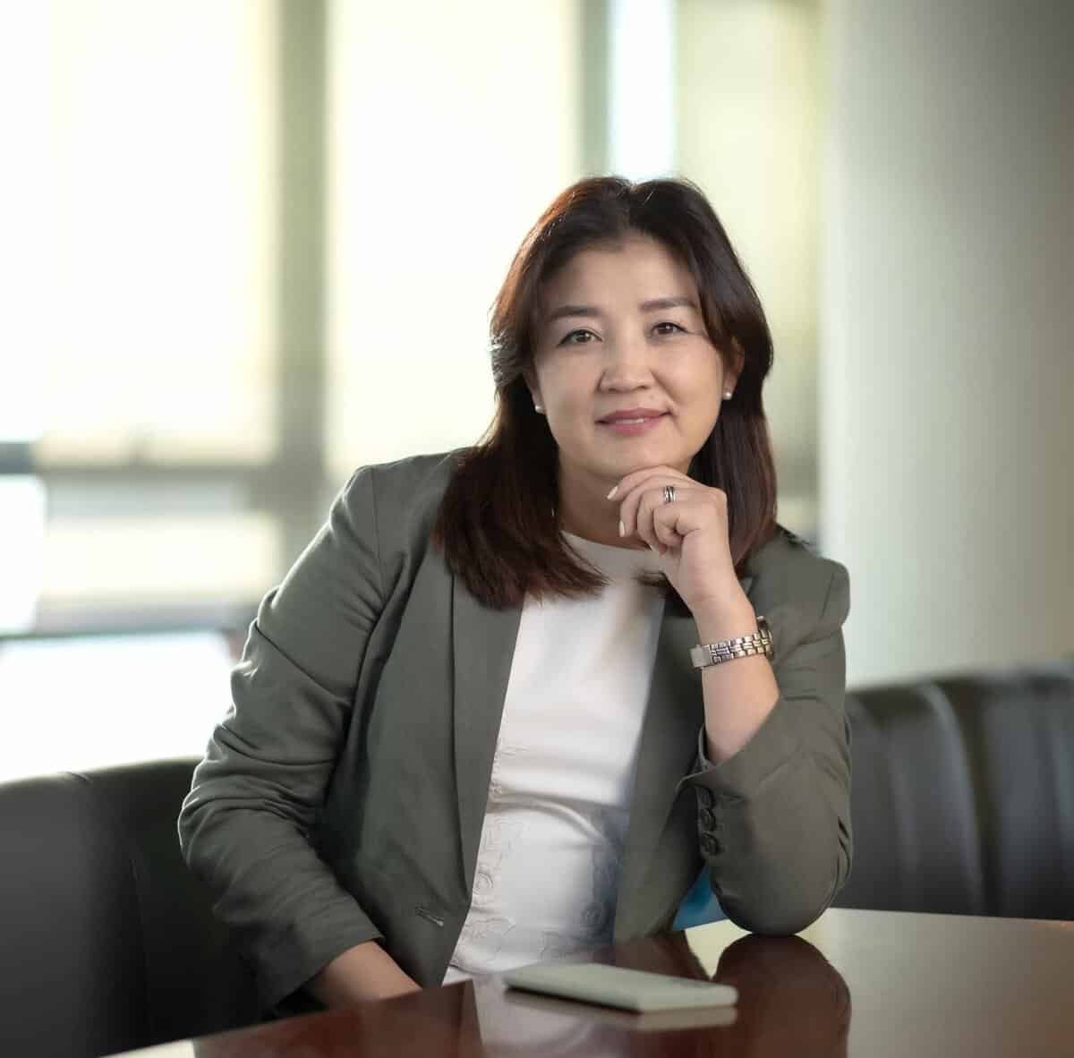 Урт хар үстэй, саарал пиджак, цагаан цамц өмссөн эмэгтэй эрүүгээ гартаа наан ширээний ард сууна. Тэр зөөлөн инээмсэглэж, камер руу шууд хардаг. Түүний урд талын ширээн дээр ухаалаг утас тавьсан нь түүний бизнесийн амбицыг харуулсан бөгөөд дэвсгэр нь зөөлөн гэрэлтдэг. - Business.MN