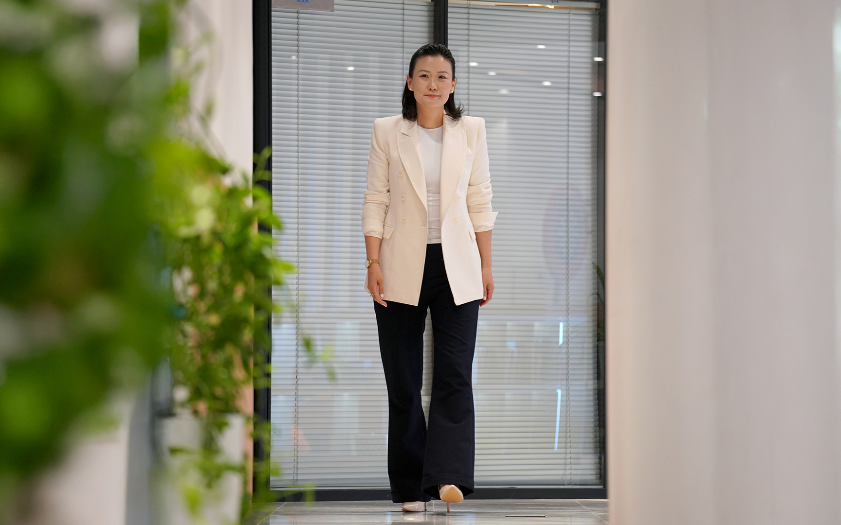 A woman wearing a white blazer and black pants walks confidently down a hallway with green plants on the side and glass doors behind her, embodying бизнес бол итгэлийн цамхаг босгох хамтын хөдөлмөр.