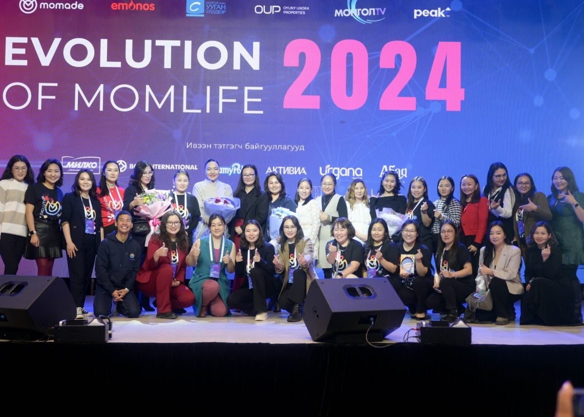 A large group of people stand on stage in front of a screen displaying "EVOLUTION OF MOMLIFE 2024." Some hold awards and bouquets. The group appears happy and celebratory.