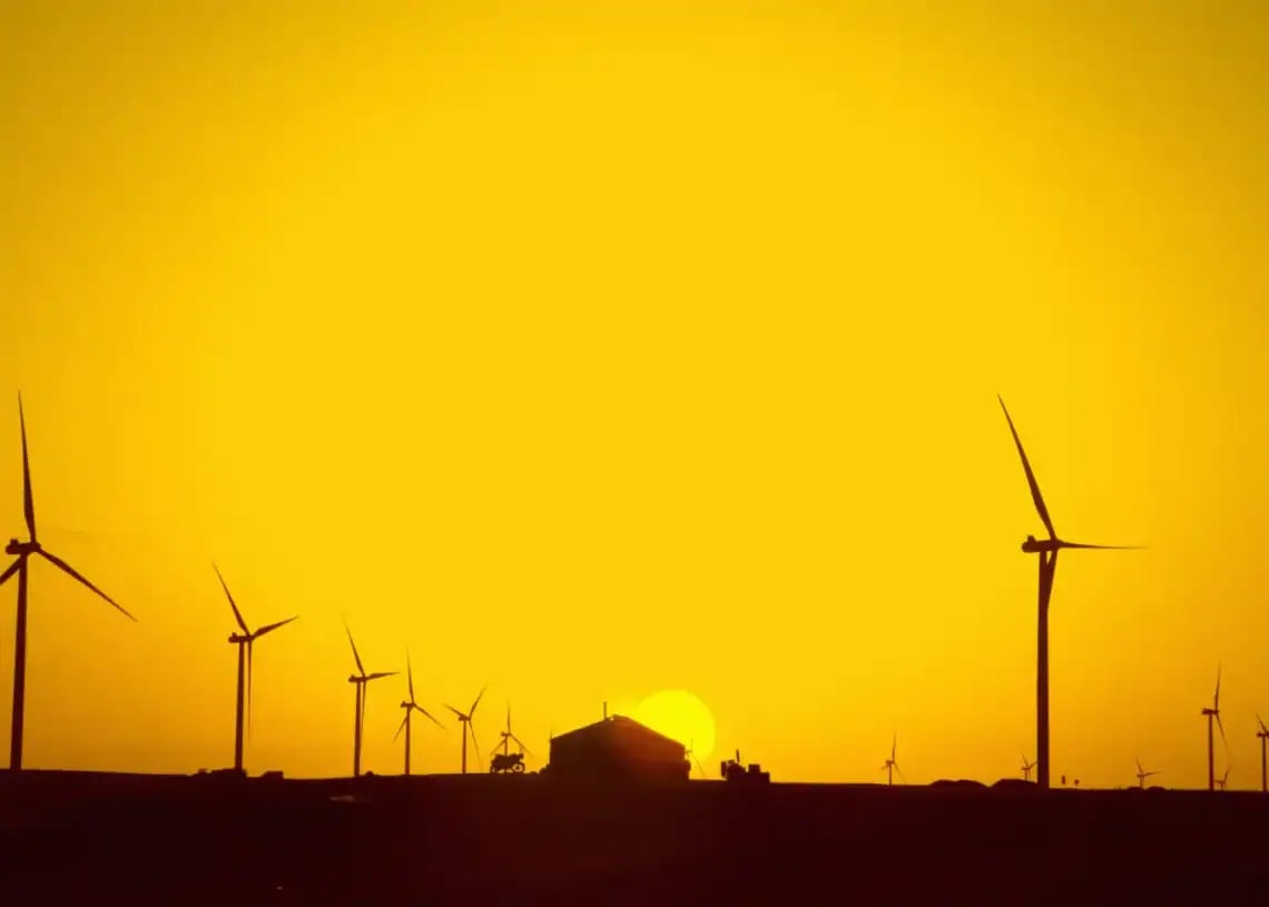 Silhouetted wind turbines and a house are seen against a vibrant orange sunset sky, with the sun sitting low on the horizon.
