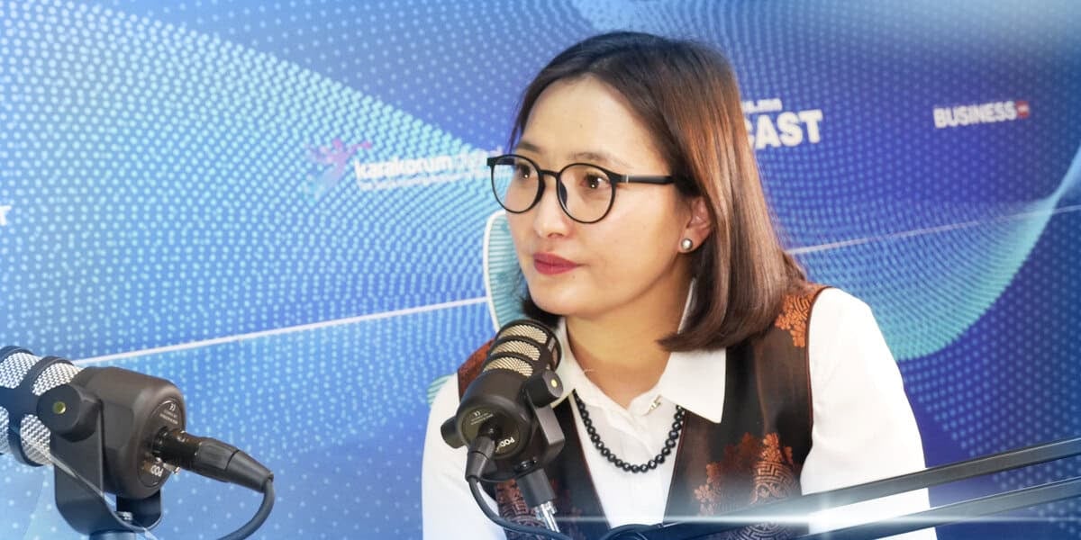 Подкаст бичлэгийн үеэр нүдний шил, хантааз өмссөн эмэгтэй микрофон руу ярьж байна. Тэр тасархай хээ, бичвэр лого бүхий цэнхэр дэвсгэр дээр сууж байна. - Business.MN