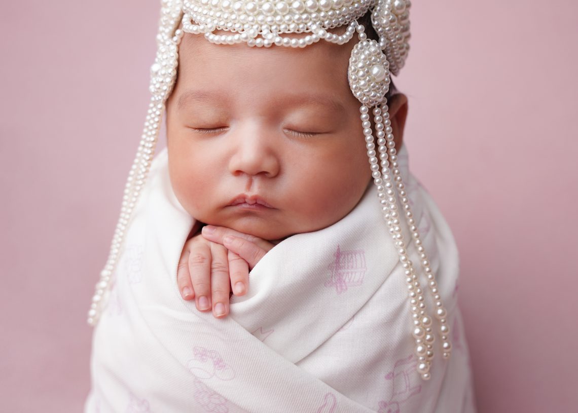 Дөнгөж төрсөн хүүхэд цайвар ягаан өнгийн хээтэй цагаан хөнжилдөө нөмрөн, зөөлөн ягаан дэвсгэр дээр хээ угалзтай сувдан малгай өмссөн гараараа эрүүгээ наан тайван унтаж байна. - Business.MN