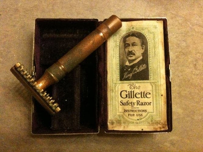 Хувцасны гуулин бариултай Gillette аюулгүй сахлын машин нь эрэгтэй хүний дүрс, "Жиллетт сахлын машин хэрэглэх заавар" гэсэн бичээстэй хайрцагны дэргэд байрладаг. Эд зүйлс нь төвийг сахисан гадаргуу дээр байна. - Business.MN