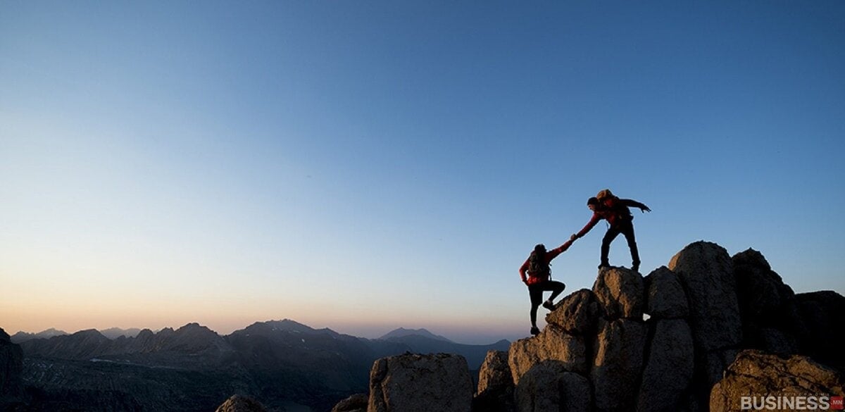 Нар ургах эсвэл жаргах үед хад чулуурхаг газар дээр дүрсэлсэн хоёр явган аялагч. Нэг хүн нөгөөдөө авирахад нь туслахаар гараа сунгадаг. Цэлмэг тэнгэрийн дор тэнгэрийн хаяанд уулс сунадаг. - Business.MN